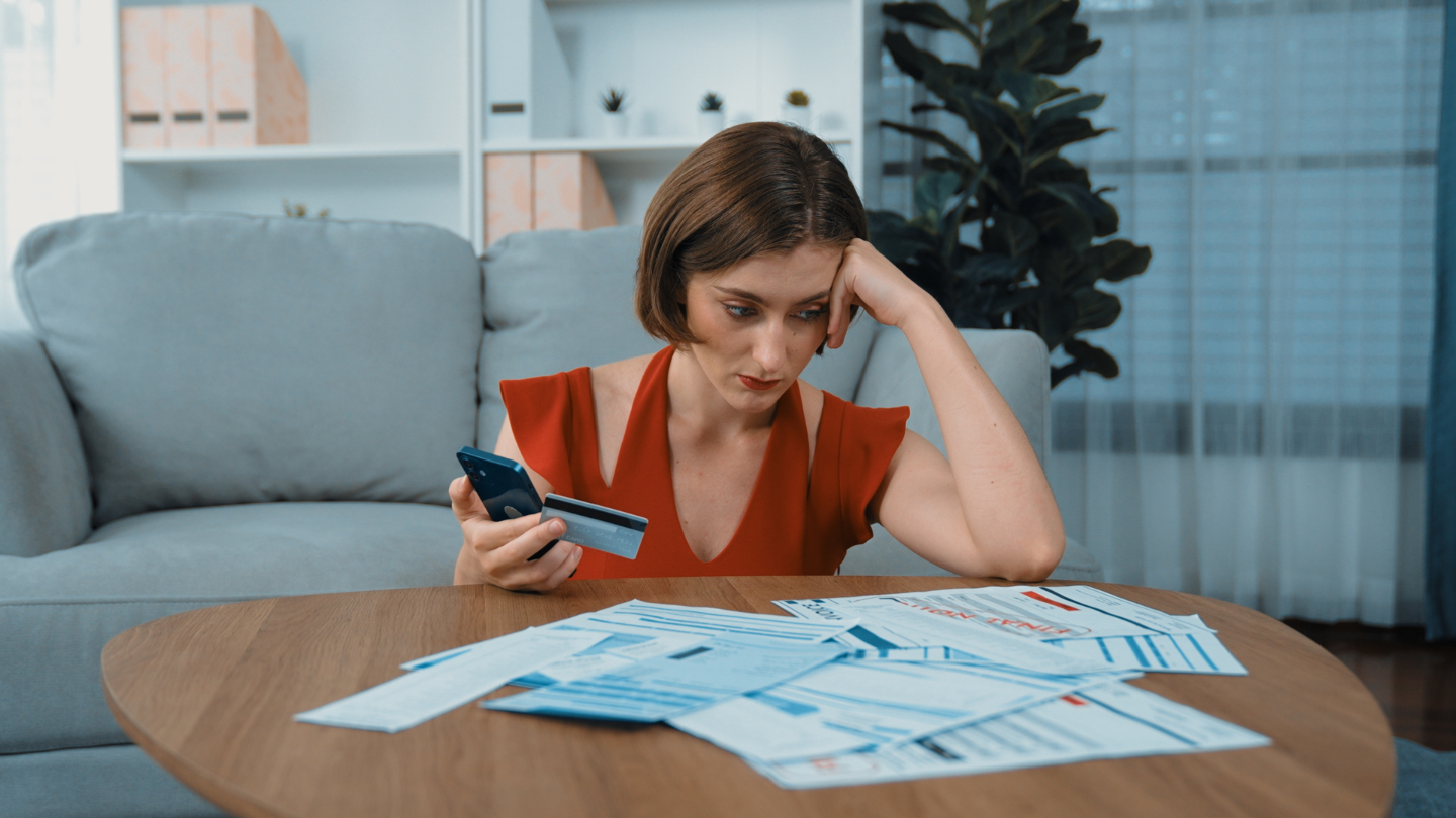 Mujer estresada mirando sus cuentas, con una tarjeta de crédito en la mano